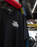 Back of the Tire Streets 'For the Streets' T-shirt hanging in a garage next to stacked tires, showing off the bold graffiti text design.