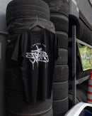 Tire Streets T-shirt displayed front-facing, hanging in a garage with tires nearby, featuring the Hand Style Tire Streets logo for a street-inspired look