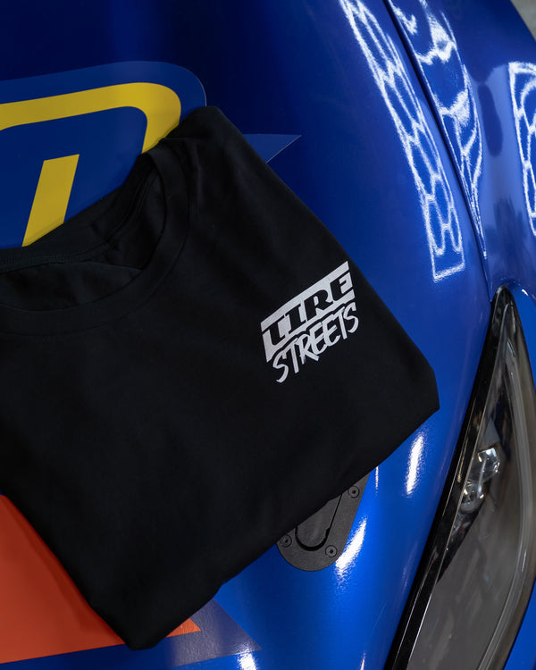 Back of the Tire Streets 'Freak in the Streets' black T-shirt featuring a tire graphic and 'Freak in the Streets' text, displayed flat on the hood of Nate Hamilton’s car