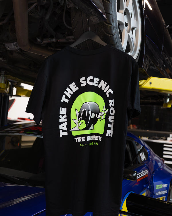 Back of the Tire Streets 'Take the Scenic Route' T-shirt featuring the wheel mascot and slogan, hanging from a car lift in a garage.