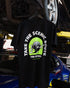 Back of the Tire Streets 'Take the Scenic Route' T-shirt featuring the wheel mascot and slogan, hanging from a car lift in a garage.