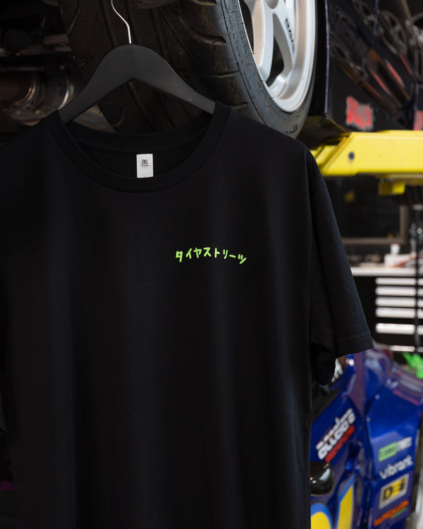 Tire Streets 'Take the Scenic Route' black T-shirt with green Japanese text hanging from a car lift in a garage.